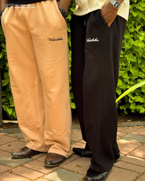 Two pairs of pants, one beige and one black, with 'Wardobe' branding, standing on a stone path with greenery in the background.