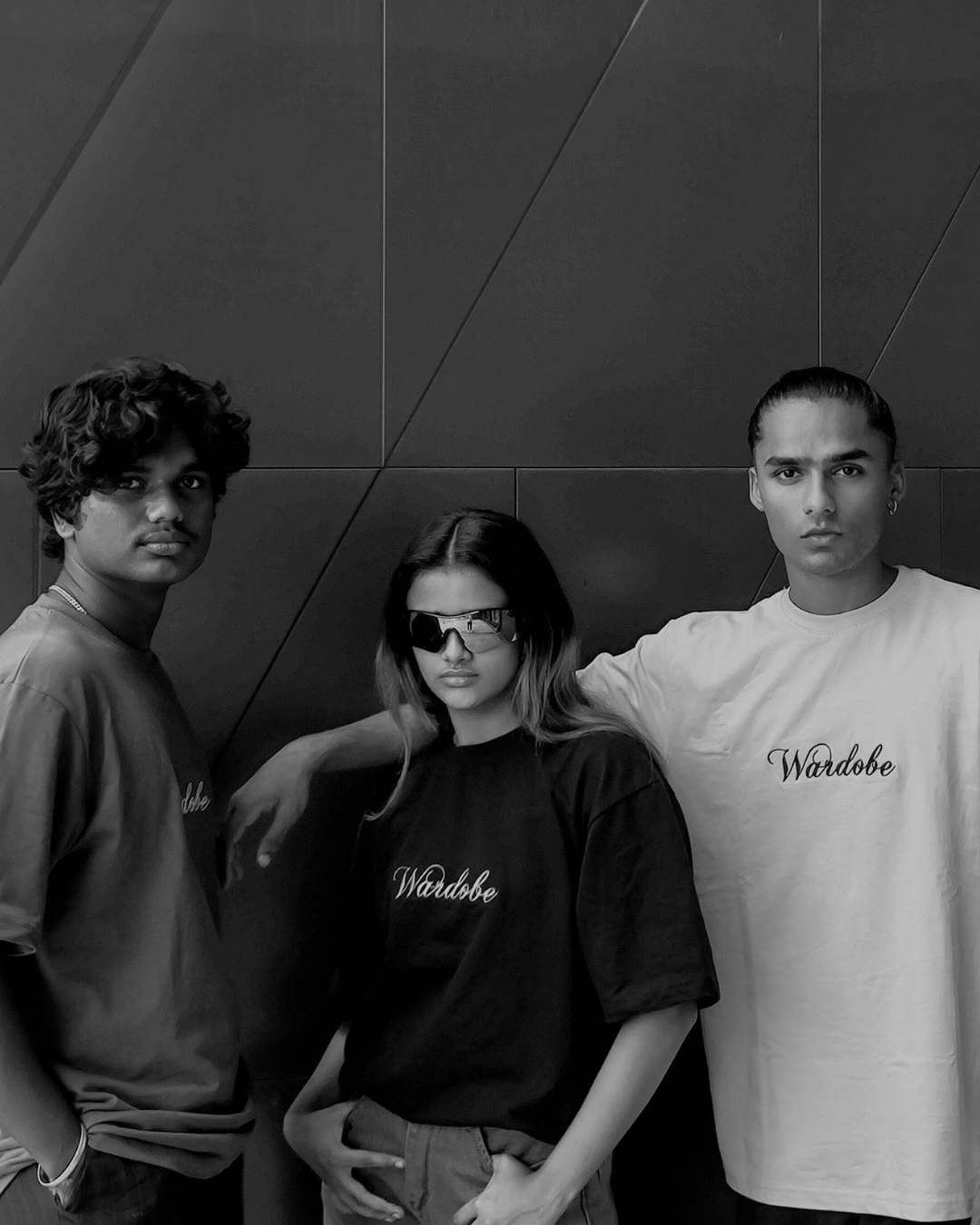 A black and white photo featuring three models from Wardobe's Embroidery Collection. The models are wearing t-shirts with an embroidered "Wardobe" logo on the chest: the male model on the left wears an olive green shirt, the female model in the center wears a black shirt, and the male model on the right wears a cream-colored shirt. They are posed in front of a dark, geometric wall.